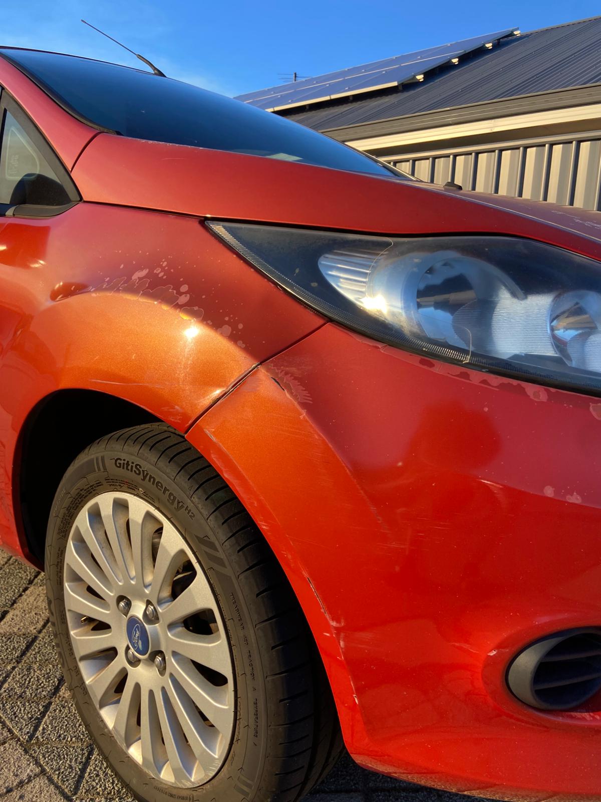 A red Ford Fiesta front guard with paint transfer scuffed along the panel beside the headlight.